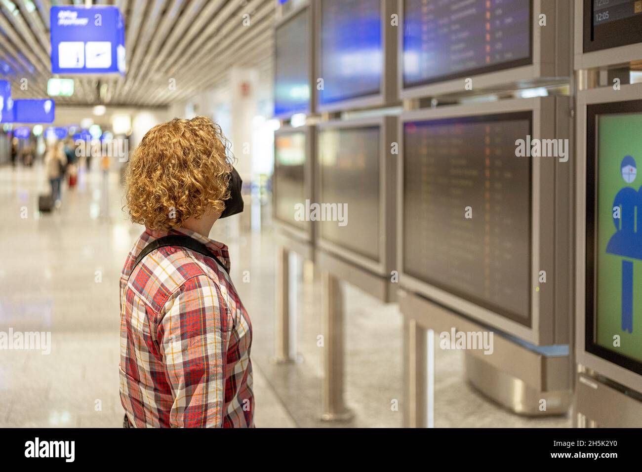 Tourist wearing an FFP2 face mask in front of the flight information boards at an airport. The