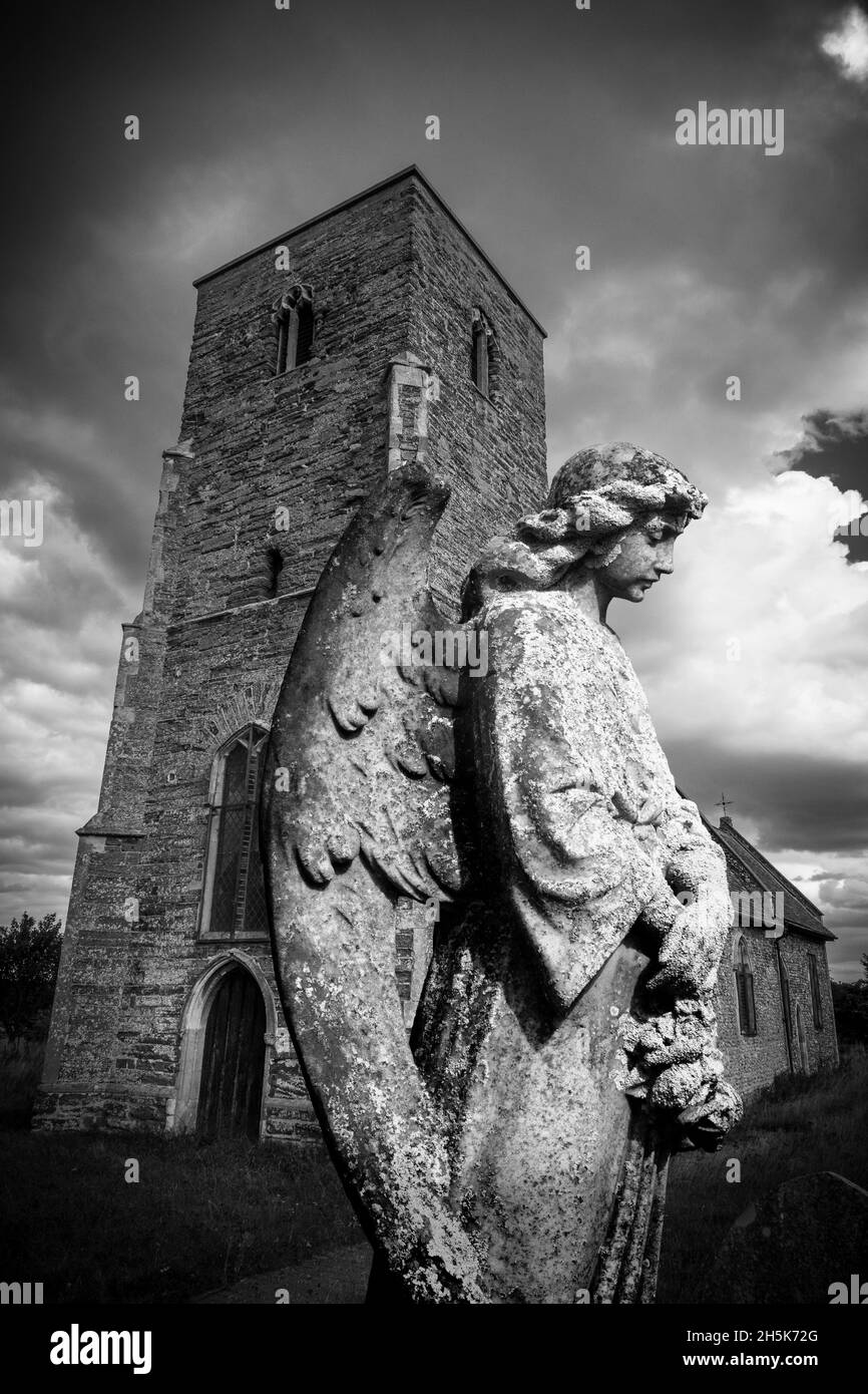 Image of a carved angel figure headstone in front of a church in a graveyard with dramatic black and white sky. Stock Photo