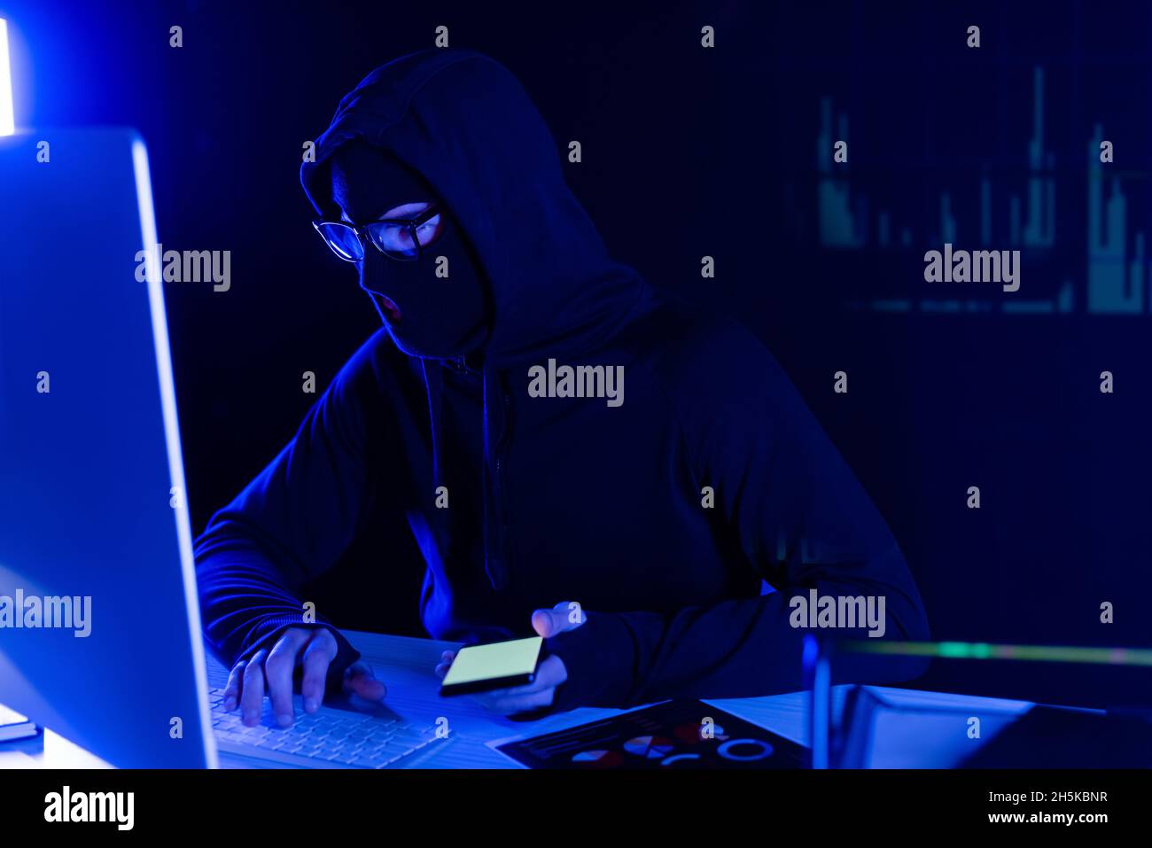 Hacker in balaclava using computer and smartphone on black background with charts reflection Stock Photo