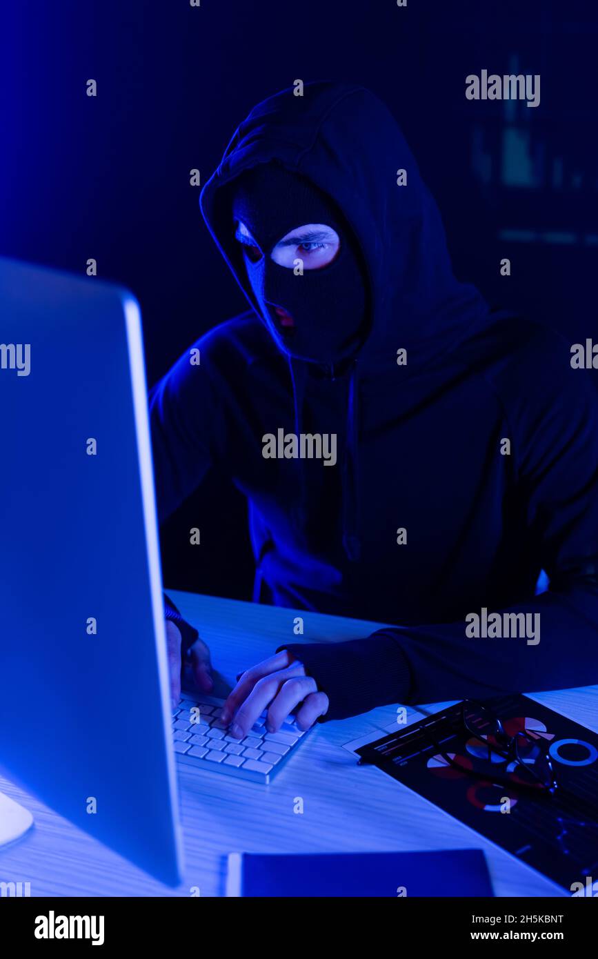 Hacker in balaclava using computer near papers on table on black background Stock Photo