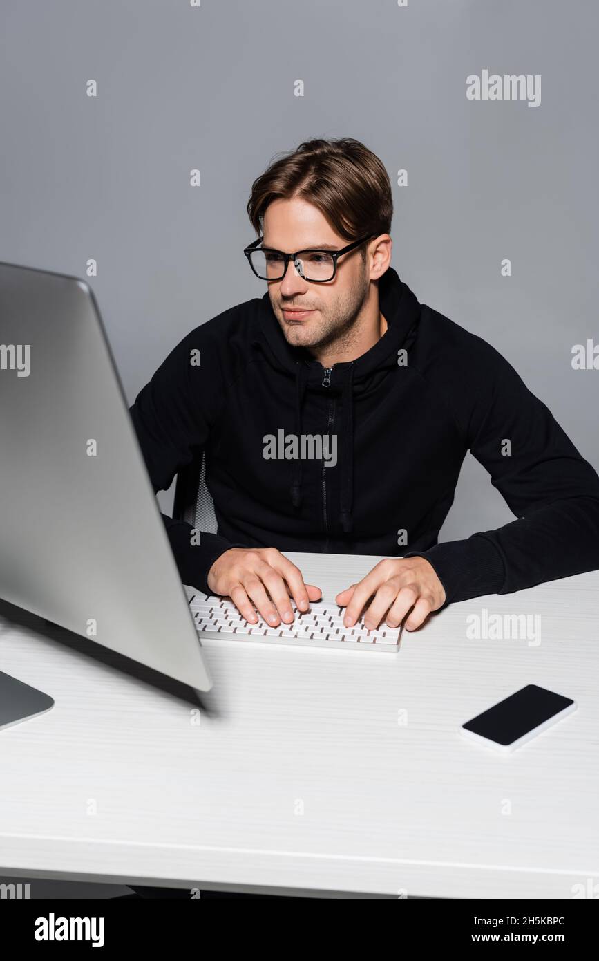 Programmer in eyeglasses using computer near smartphone isolated on grey Stock Photo