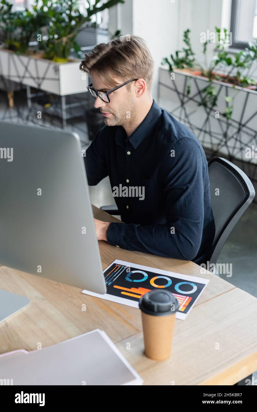 Programmer in eyeglasses using computer near document and coffee in office Stock Photo