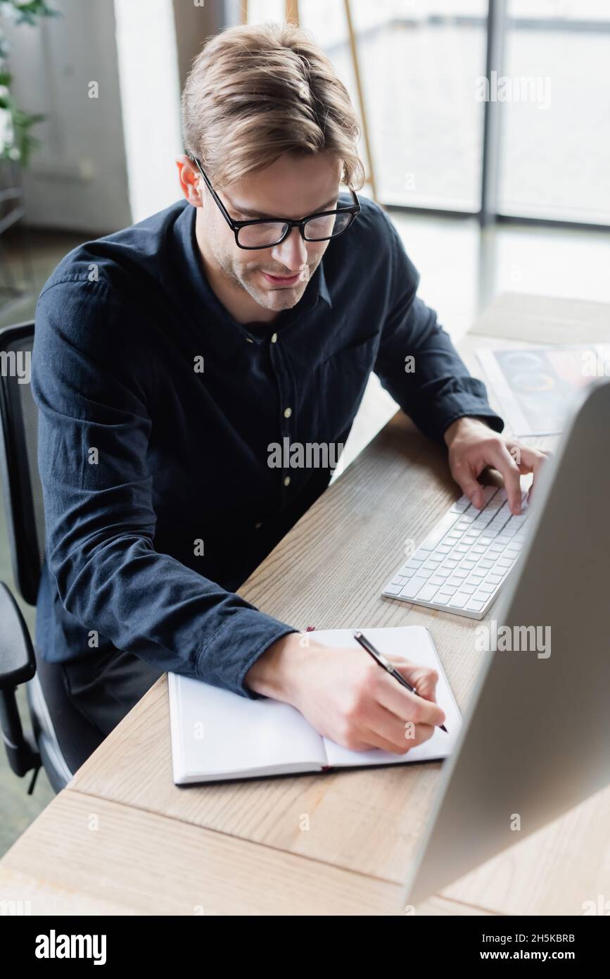 Programmer in eyeglasses writing on notebook and using computer in office Stock Photo