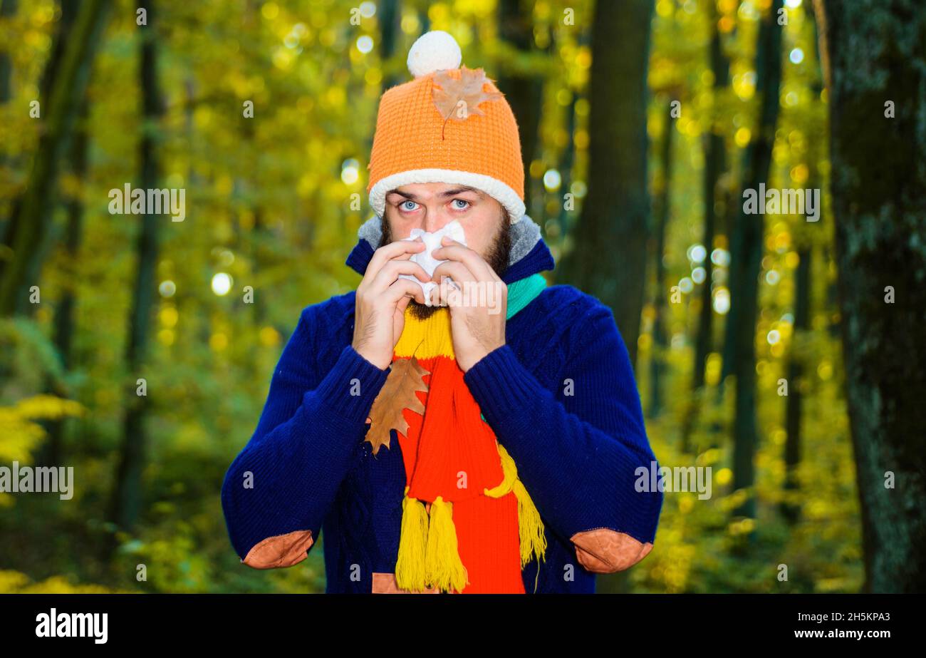 Sick man in autumn park sneezing in napkin. Guy coughing. Influenza, illness, sick, cold. Coronavirus epidemic. Male having runny nose. Stock Photo