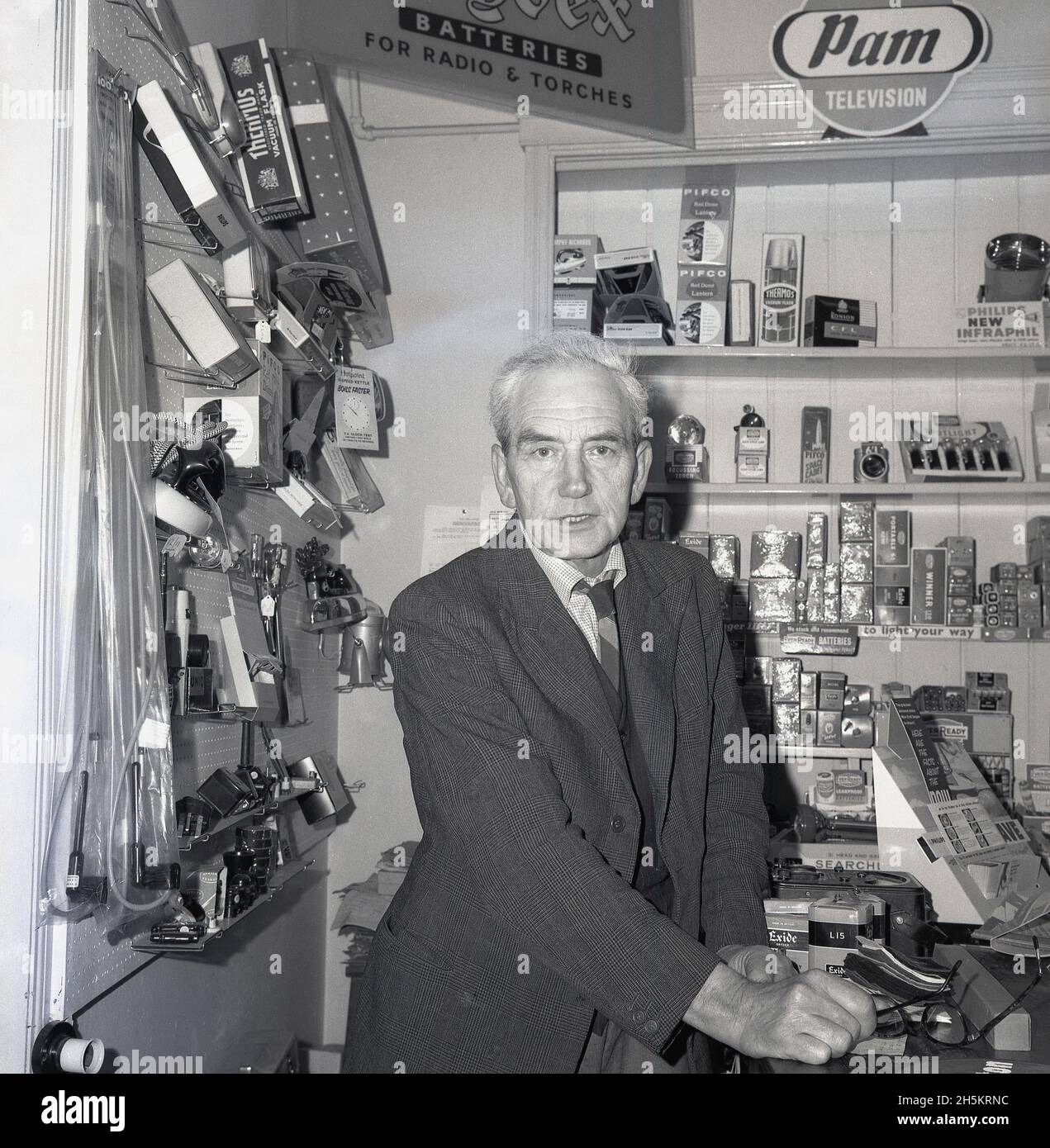 1960, a male shopkeeper at the counter of his electrical goods store, England, UK. Behind his right shoulder on the wall a traditional peg board, used to display smaller items; irons, lights, clocks, torches, plugs. On wooden shelves, a promotional card for Pam, a brand name of British manufacturer Pamphonic. Founded by Paul Taylor and Edgar Lavington in 1932 in the garage of his mother's house in Prince Albert St, London, Pamphonoc Reproducers Ltd made high quality sound systems. 1n 1947 Pye brought control. Stock Photo