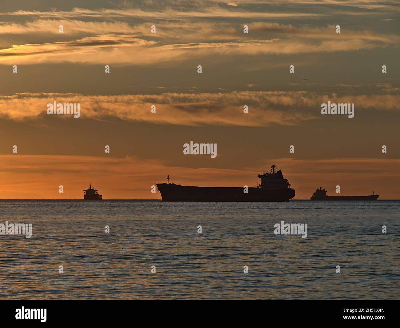 Silhouettes of three cargo freight vessels mooring in English Bay in front of Vancouver, British Columbia, Canada at sunset with orange dramatic sky. Stock Photo