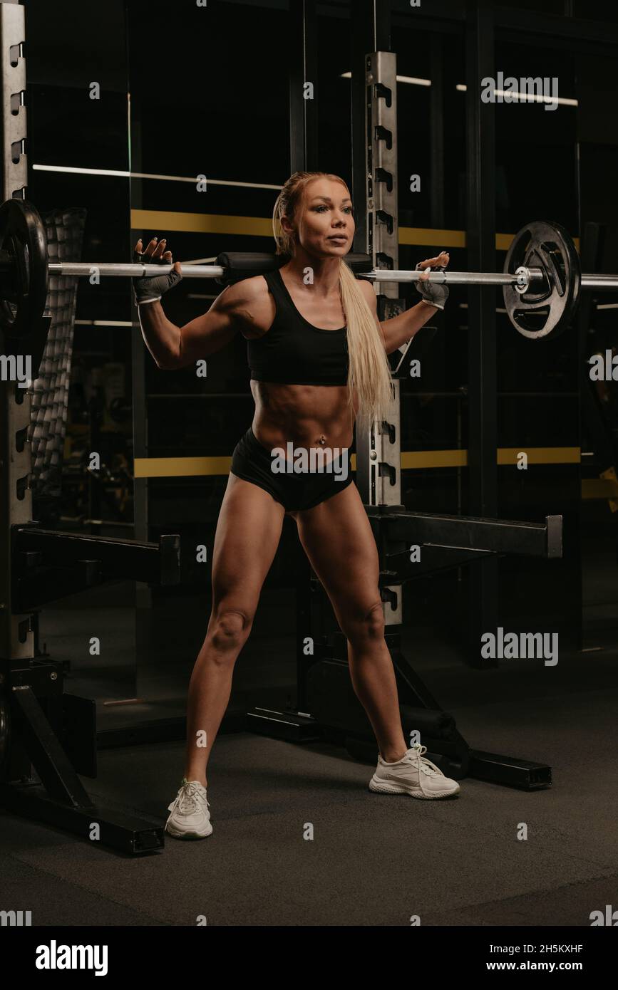 A sporty woman with blonde hair is squatting with a barbell near the squat rack in a gym. A girl is doing a leg workout Stock Photo Alamy