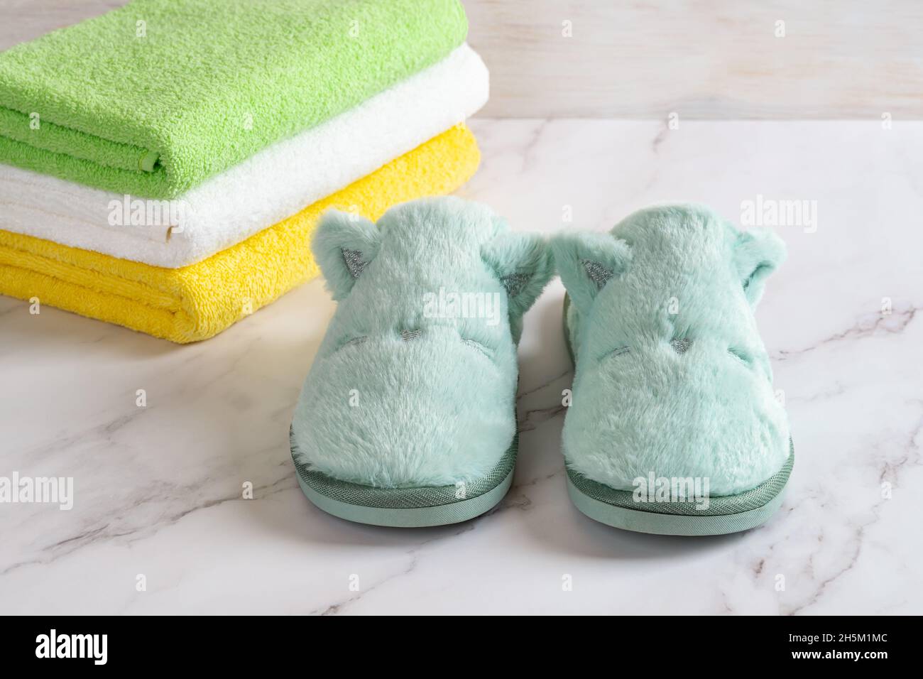 Funny cozy slippers near stack of colored terry towels over marble surface. Concepts of natural cotton toiletries, soft fleece shoes for home. Stock Photo