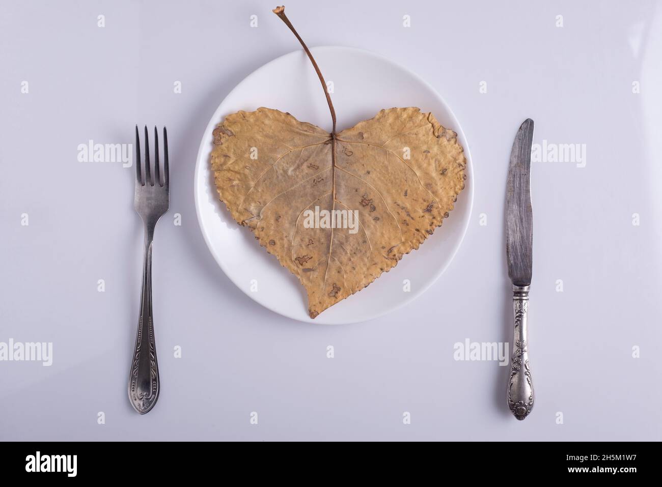 creative autumn concept heart shaped leaf on plate with cutlery cutlery on white background Stock Photo