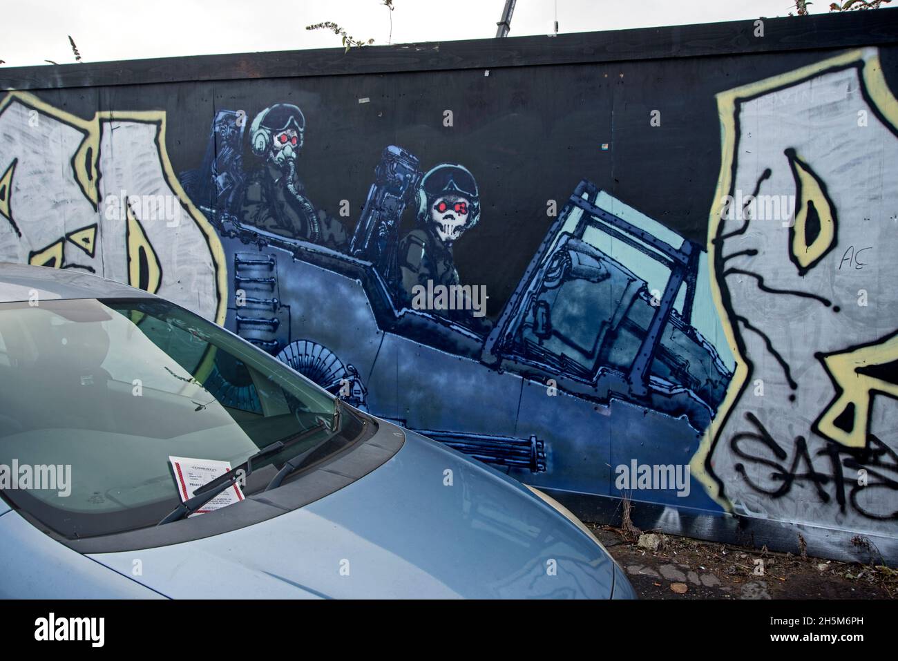 Car with parking ticket parked next to a piece of streetart featuring an aircraft with skeleton pilots. Stock Photo