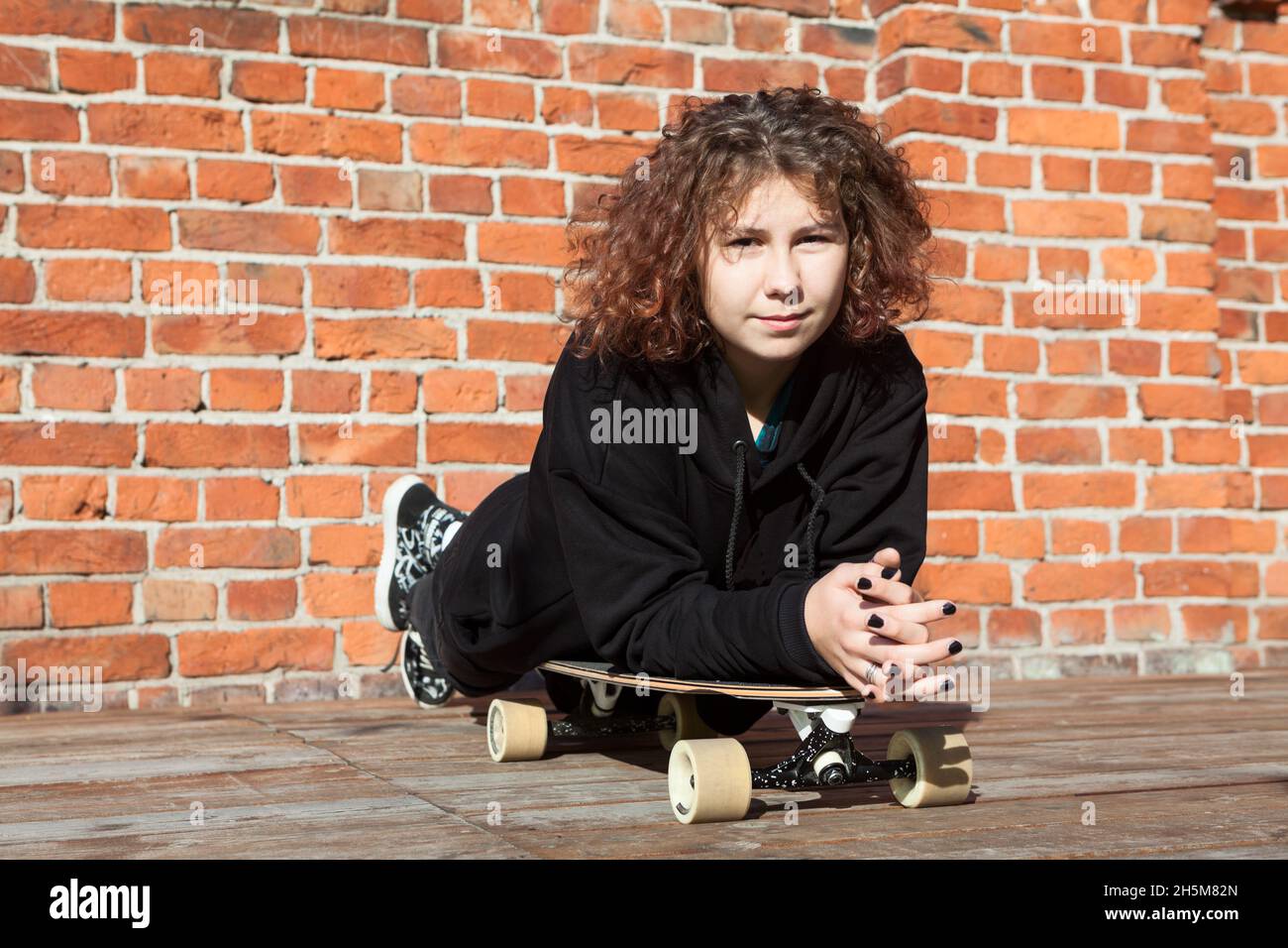 Teenage Caucasian girl lying on longboard against red brick wall
