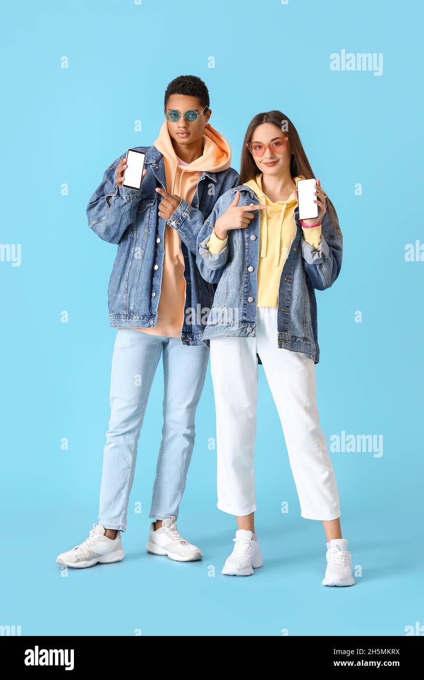 Stylish young couple in hoodies pointing at mobile phones on blue background Stock Photo