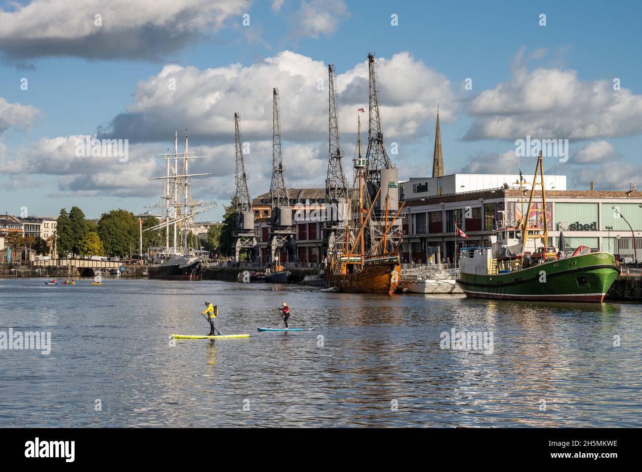 Paddleboarders paddle across Bristol's Floating Harbour, with historic ships docked outside the M Shed museum behind. Stock Photo