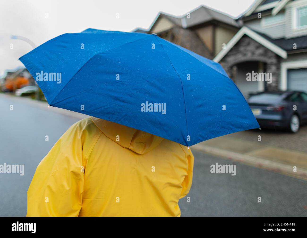 Woman with blue umbrella walking in the rain on a street. Unrecognizable person in yellow rain jacket with umbrella. Stock Photo