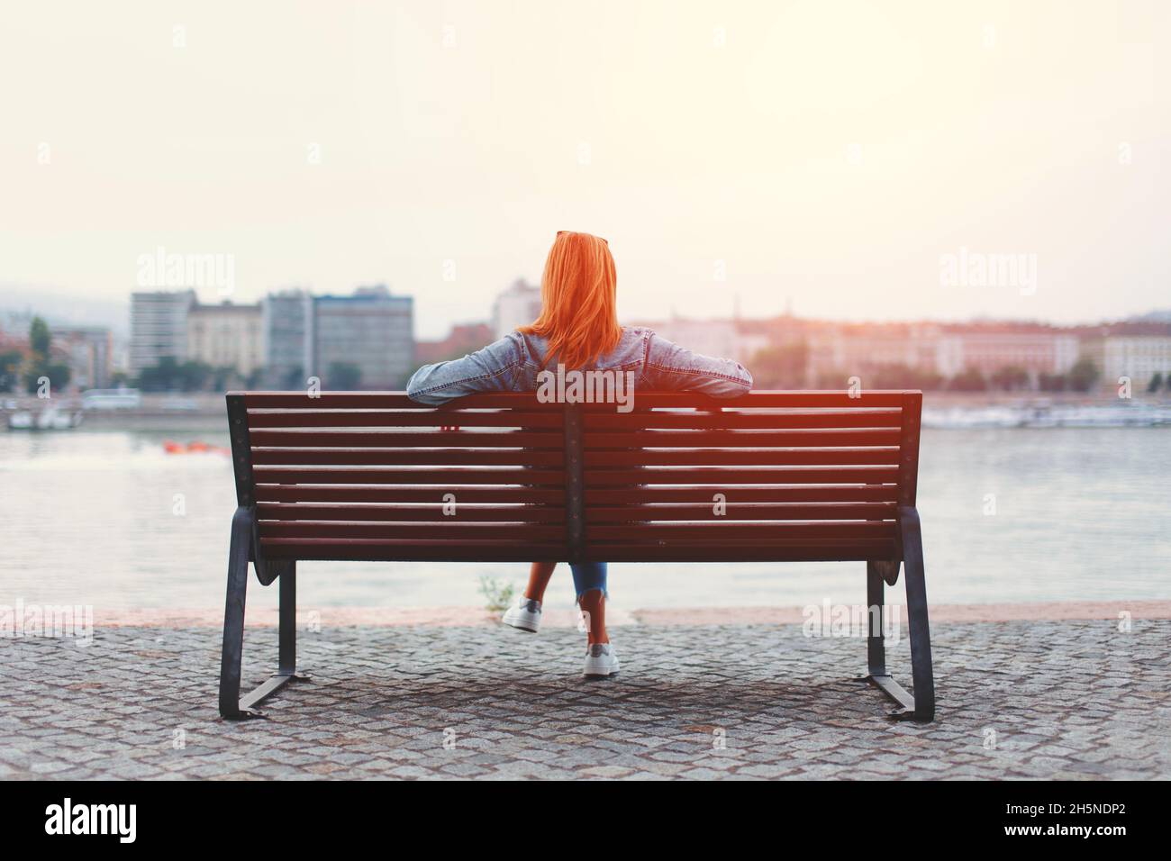 Young redhead woman relaxing on bench at riverside in sunlight Stock Photo
