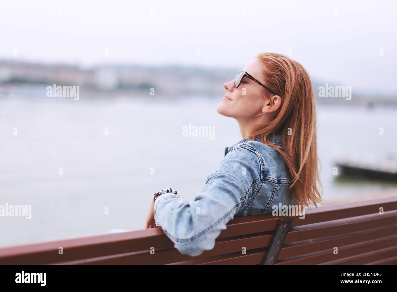 Young balanced redhead Caucasian woman sitting on bench at riverside meditating Stock Photo