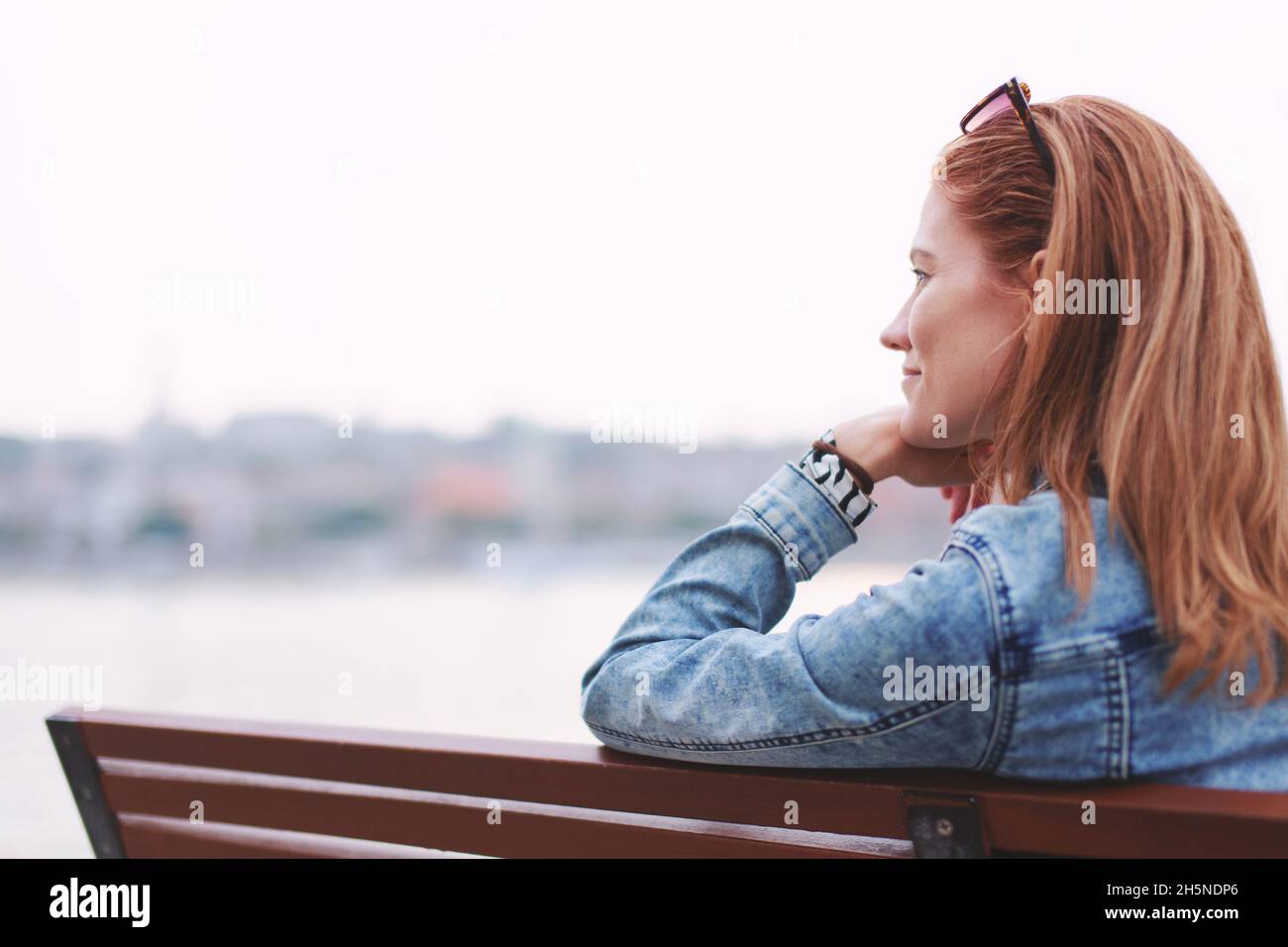 Young balanced redhead Caucasian woman sitting on bench relaxing at riverside Stock Photo