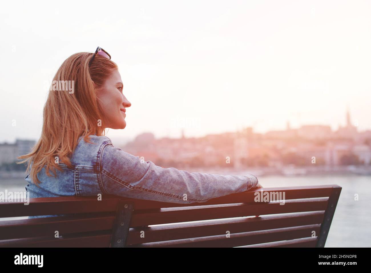 Young balanced redhead Caucasian woman sitting on bench resting at riverside in sunset Stock Photo