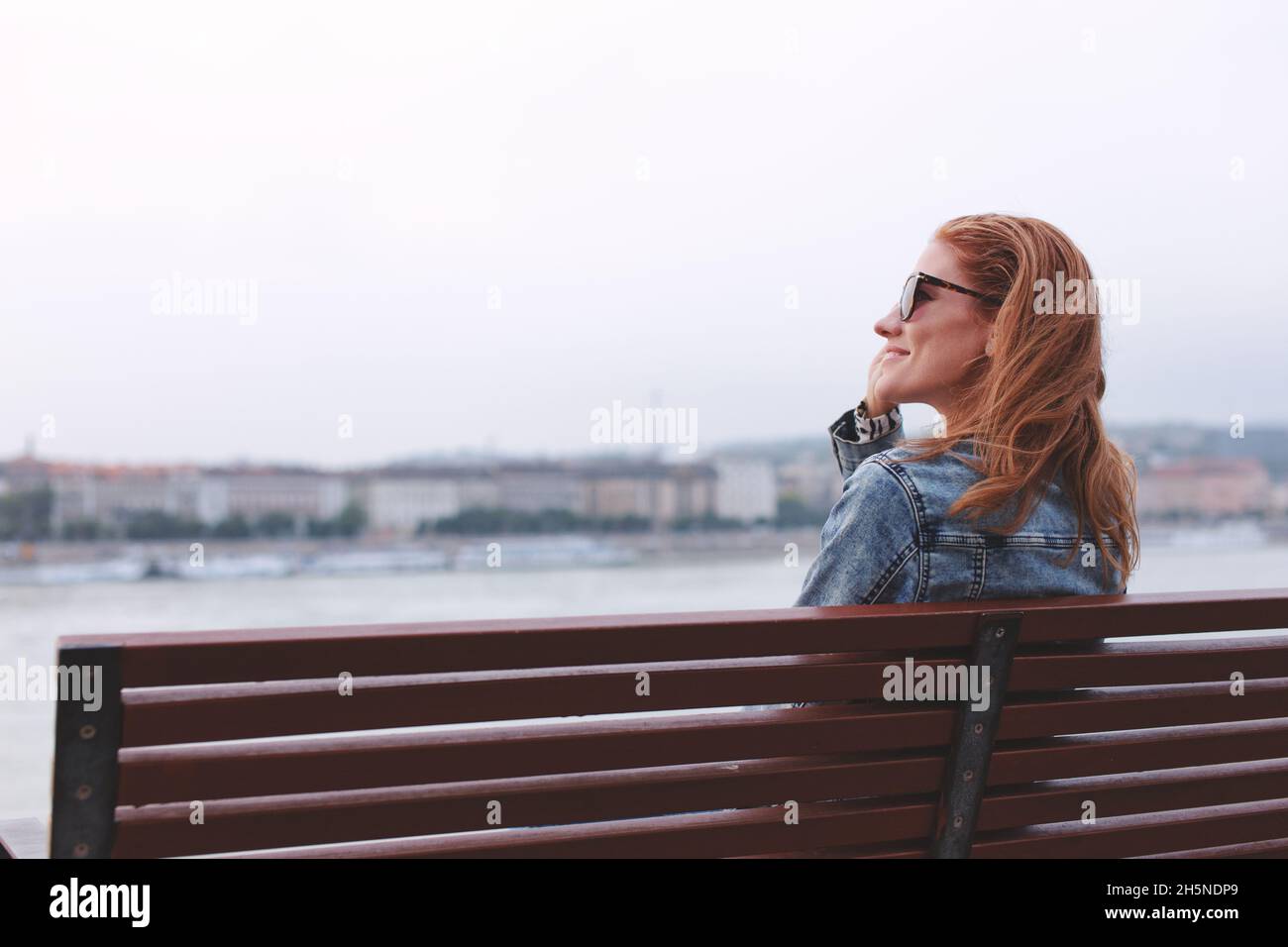 Young balanced redhead Caucasian woman sitting on bench and holding sunglasses at riverside Stock Photo