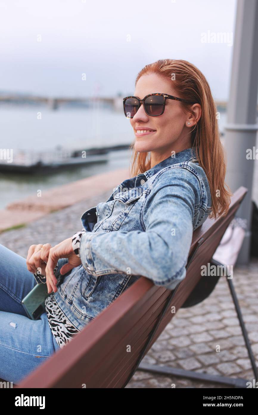 Young balanced redhead Caucasian woman sitting on bench in sunglasses at riverbank Stock Photo