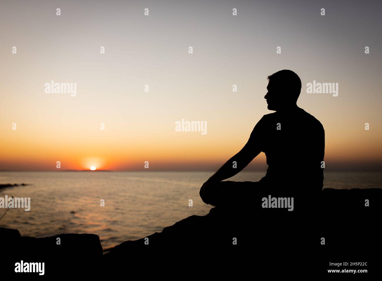 A man enjoys the view of the sunset on the sea, sitting on a rock. Side view, silhouette. Stock Photo