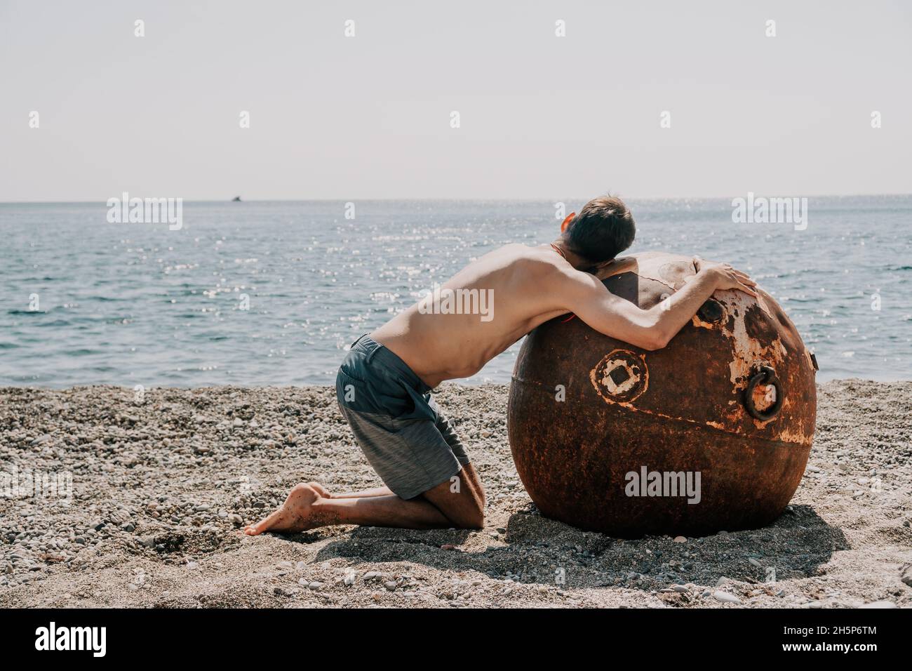 Man doing yoga and meditation outdoors near to old rusty floating marine mine on the beach with rocky shore and sea background. Healthy lifestyle Stock Photo