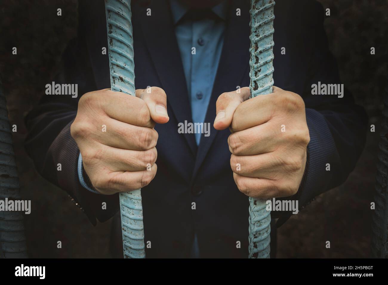 Young corrupted businessman in jail, behind the prison bars. Men's hands hold a reinforced grille in the dark. The concept of financial crimes Stock Photo