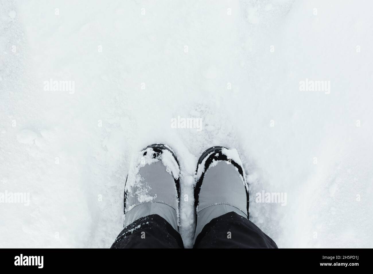 Feet snow top view. Winter gray boots stand on white snow. Textured background. Women's suit black pants and boots for walking, hiking, trekking. The Stock Photo