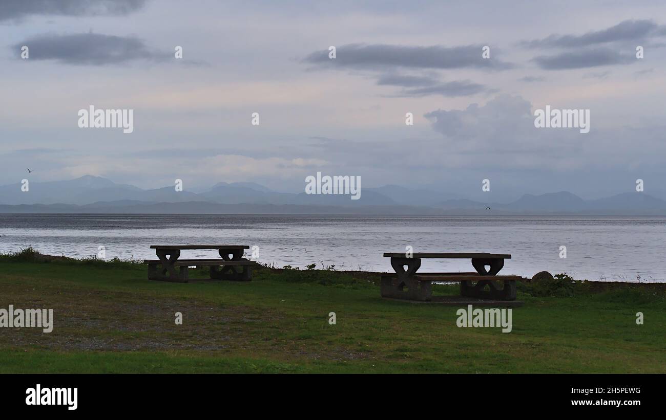Two lonely rest benches on the coast of the Pacific Ocean in Port Hardy, Vancouver Island, British Columbia, Canada with Coast Mountains on horizon. Stock Photo