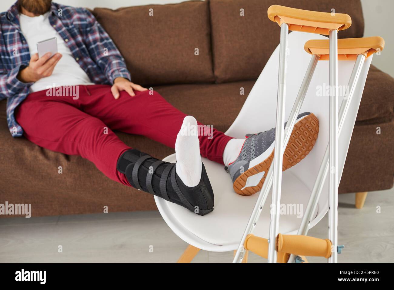 Young man with broken leg or injured foot sitting on couch and using mobile phone Stock Photo