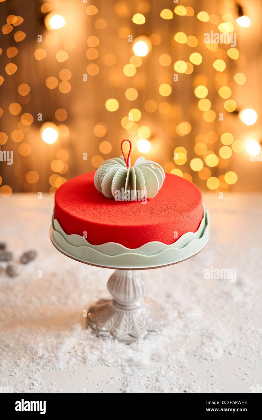 Mousse cake covered with red velour. Garland lamps bokeh on background. Modern european cake. French cuisine. Christmas theme. Stock Photo
