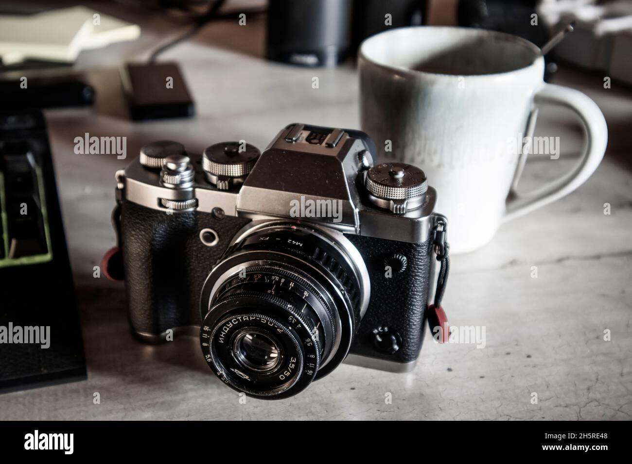 Vintage old 50mm USSR lens on a silver and black hybrid camera body on a grey desk top with coffee mug Stock Photo