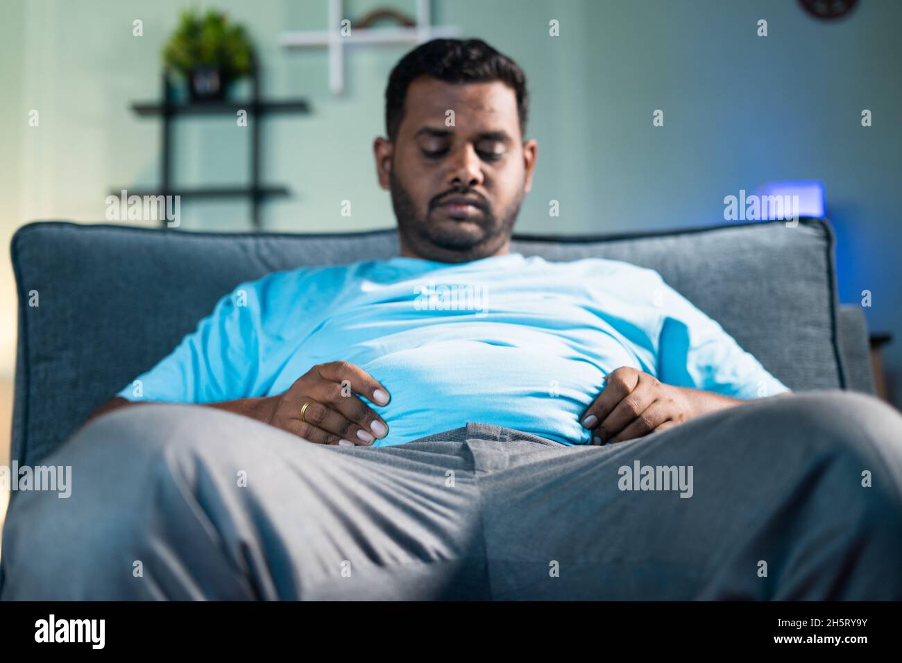 Sad obase man disappointed for heavy belly fat by holding hands while sitting on sofa at home - Concept of overweight, desease, suffering from excess Stock Photo