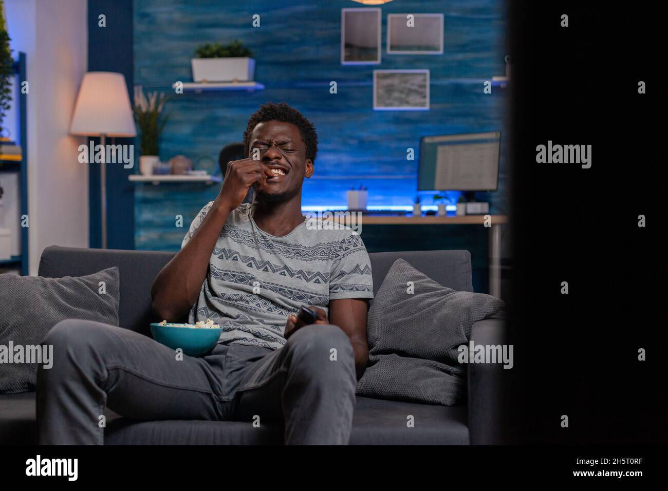 African american young man watching television laughing during comedy movie relaxing in living room. Black guy sitting on sofa while eating popcorn enjoying spending tine alone Stock Photo