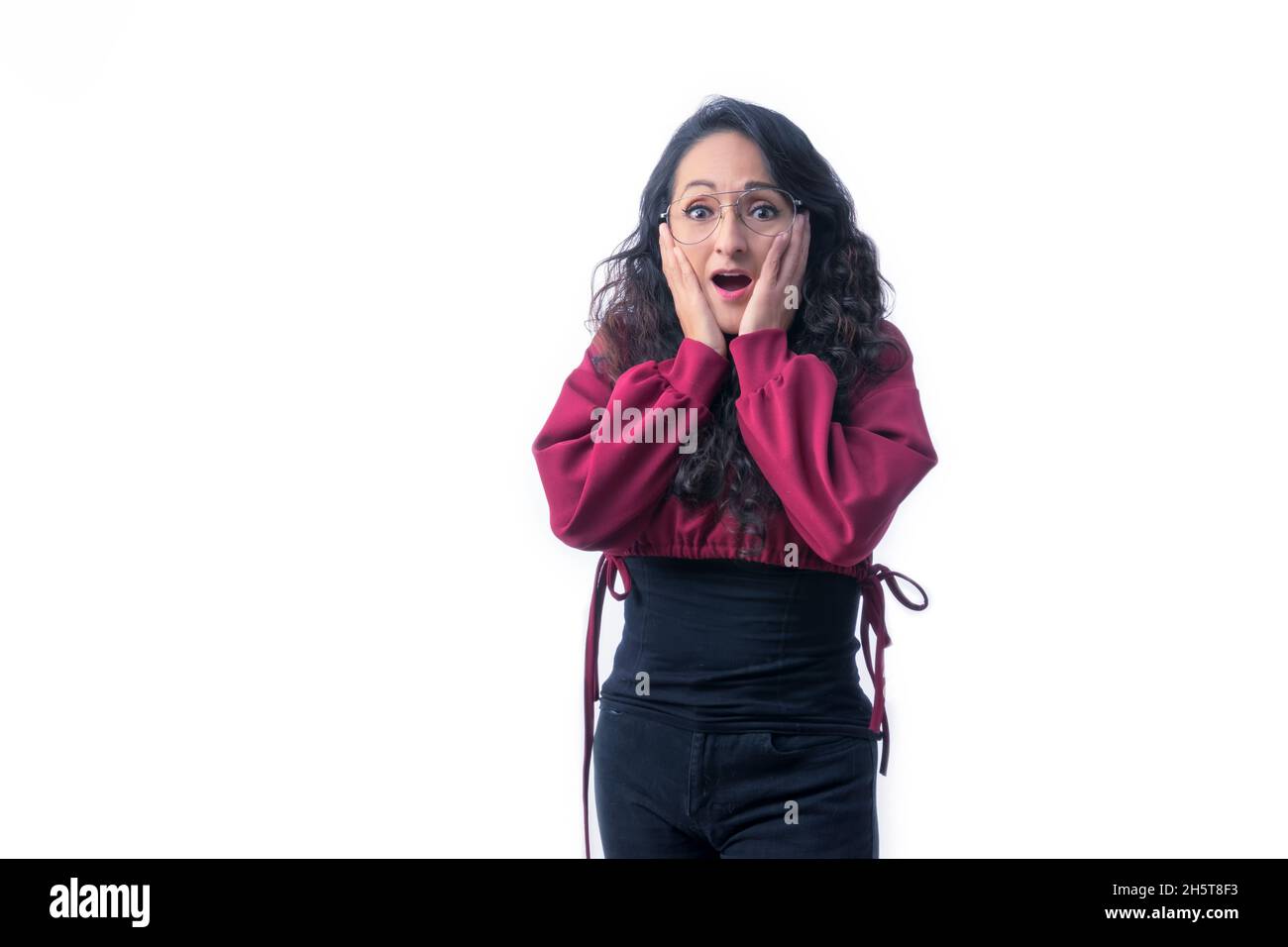Pretty Latin Hispanic woman with long thick hair wearing glasses on white background indoors making expressions and gestures, surprised, amazed, with Stock Photo