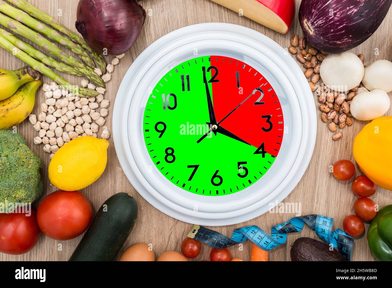 Clock, surrounded by healthy food, with one portion marked in red and one in green. Concept of intermittent fasting, a diet that provides benefits suc Stock Photo