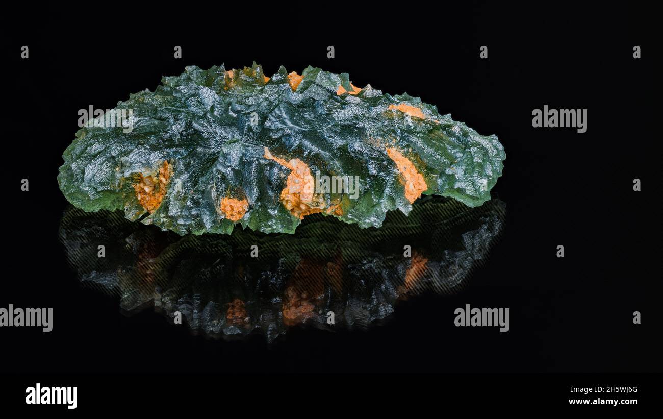 Natural green moldavite gem with reflection on a black background. Beautiful meteoric glass with orange dirt in wavy surface in artistic still life. Stock Photo