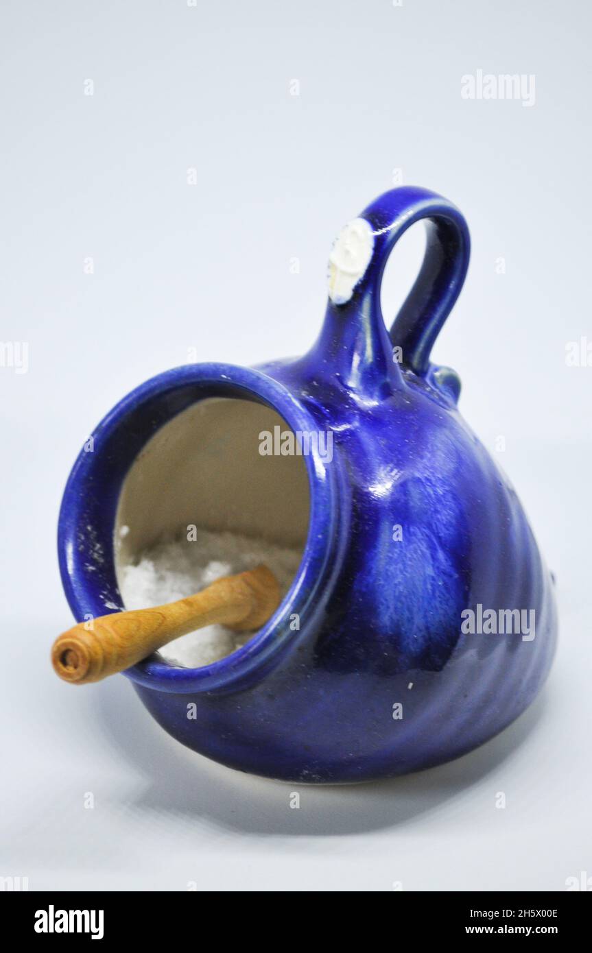 A handmade, pottery salt pig in a royal blue glaze. It is filled with salt crystals and has a small wooden spoon. It is set against a white background Stock Photo