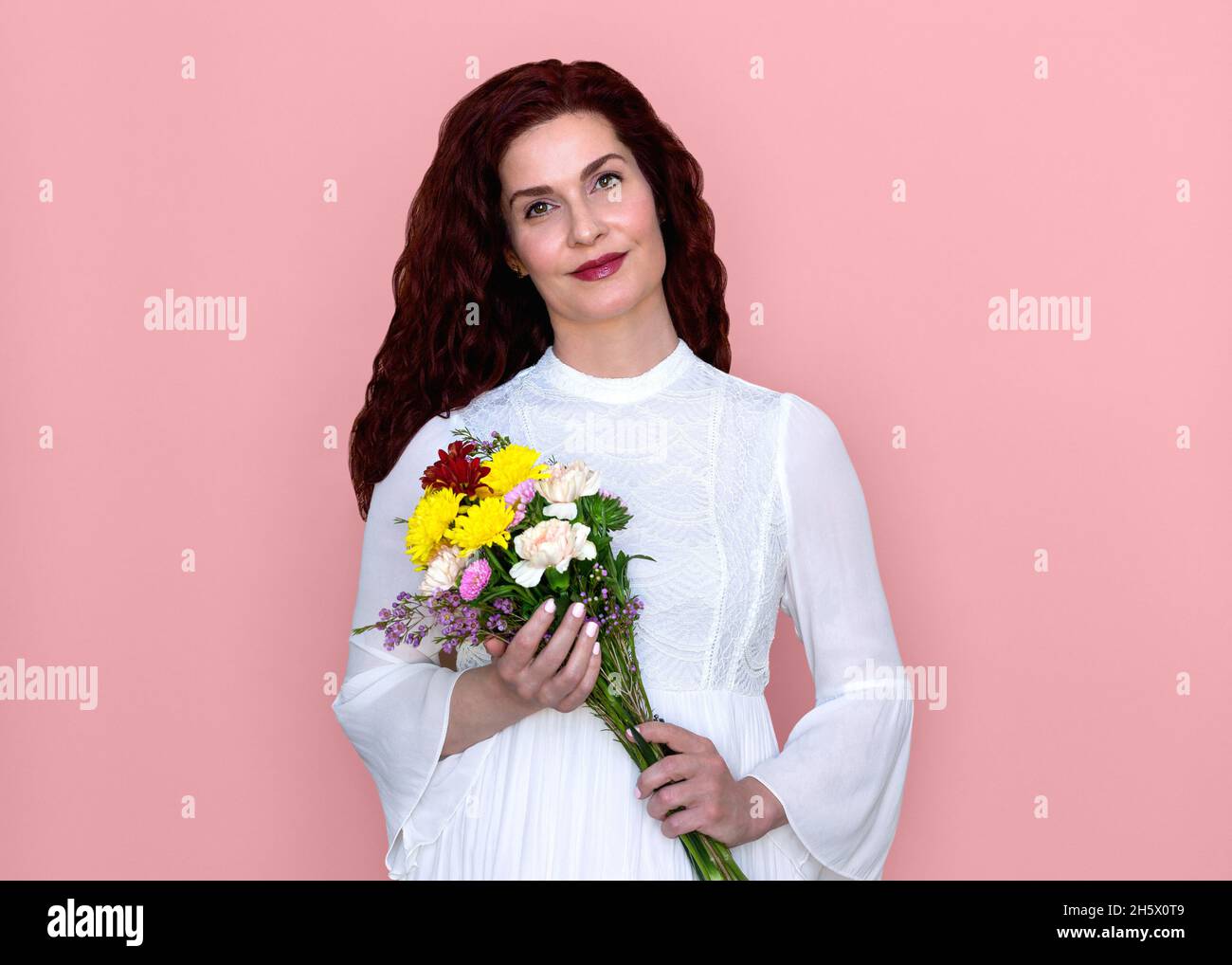 Woman Holding Flowers with Soft Smile on Pink Background. Romantic portrait of pretty woman in white dress holding bouquet of flowers across her body. Stock Photo