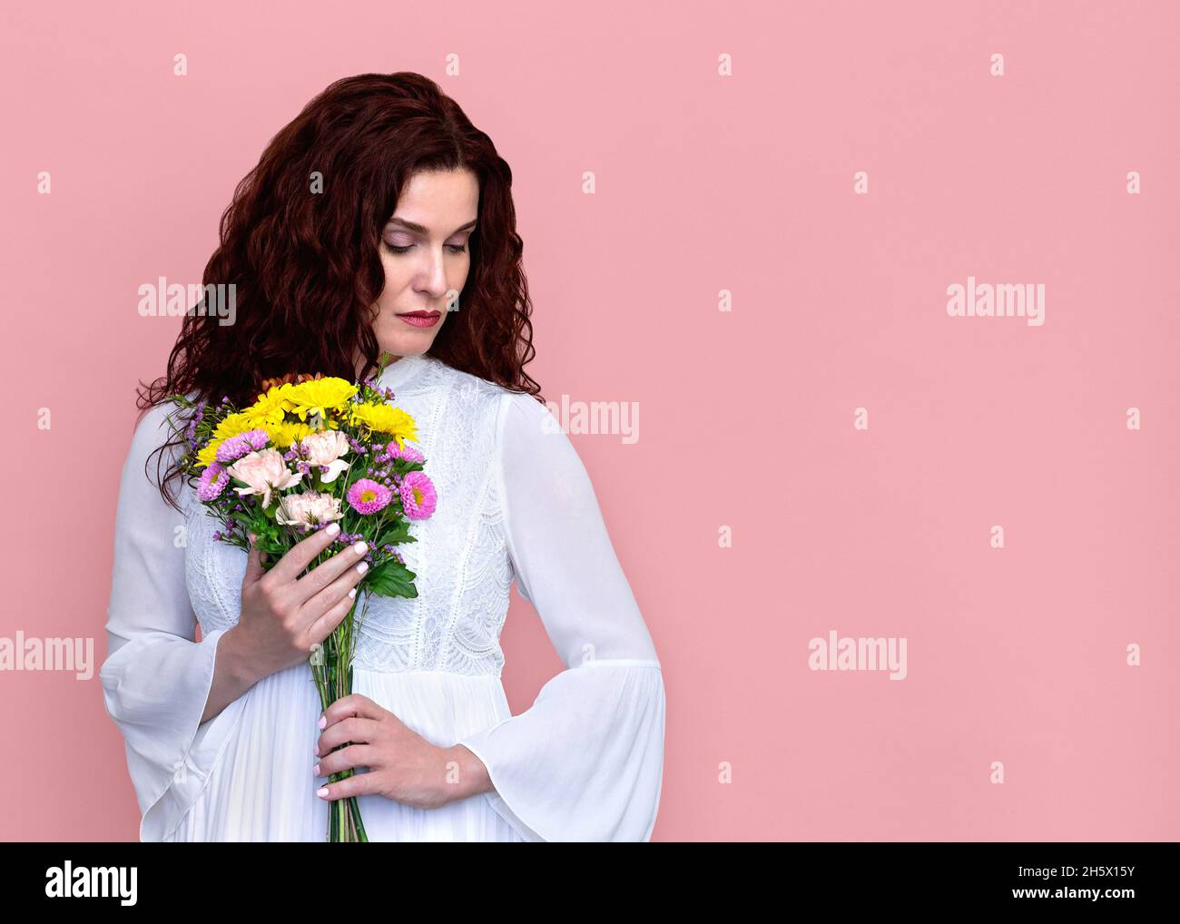 Woman Holding Flowers to Chest Looking Down on Pink Background. Portrait of sad pensive woman in white dress holding bouquet to her chest. Stock Photo