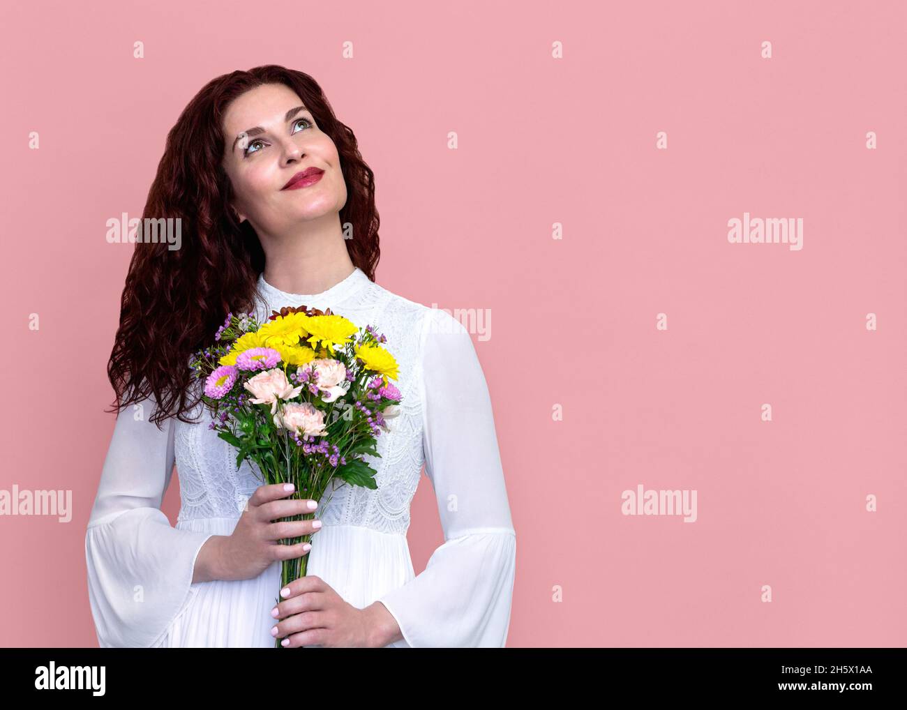 Woman Holding Flowers Looking Up with Soft Smile on Pink Background. Close-up portrait of beautiful happy woman in white dress holding bouquet. Stock Photo
