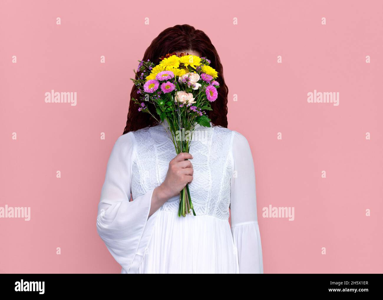 Woman with Flower Bouquet in Front of Face on Pink Background. Studio portrait of playful woman holding flower bouquet in front of her face. Stock Photo