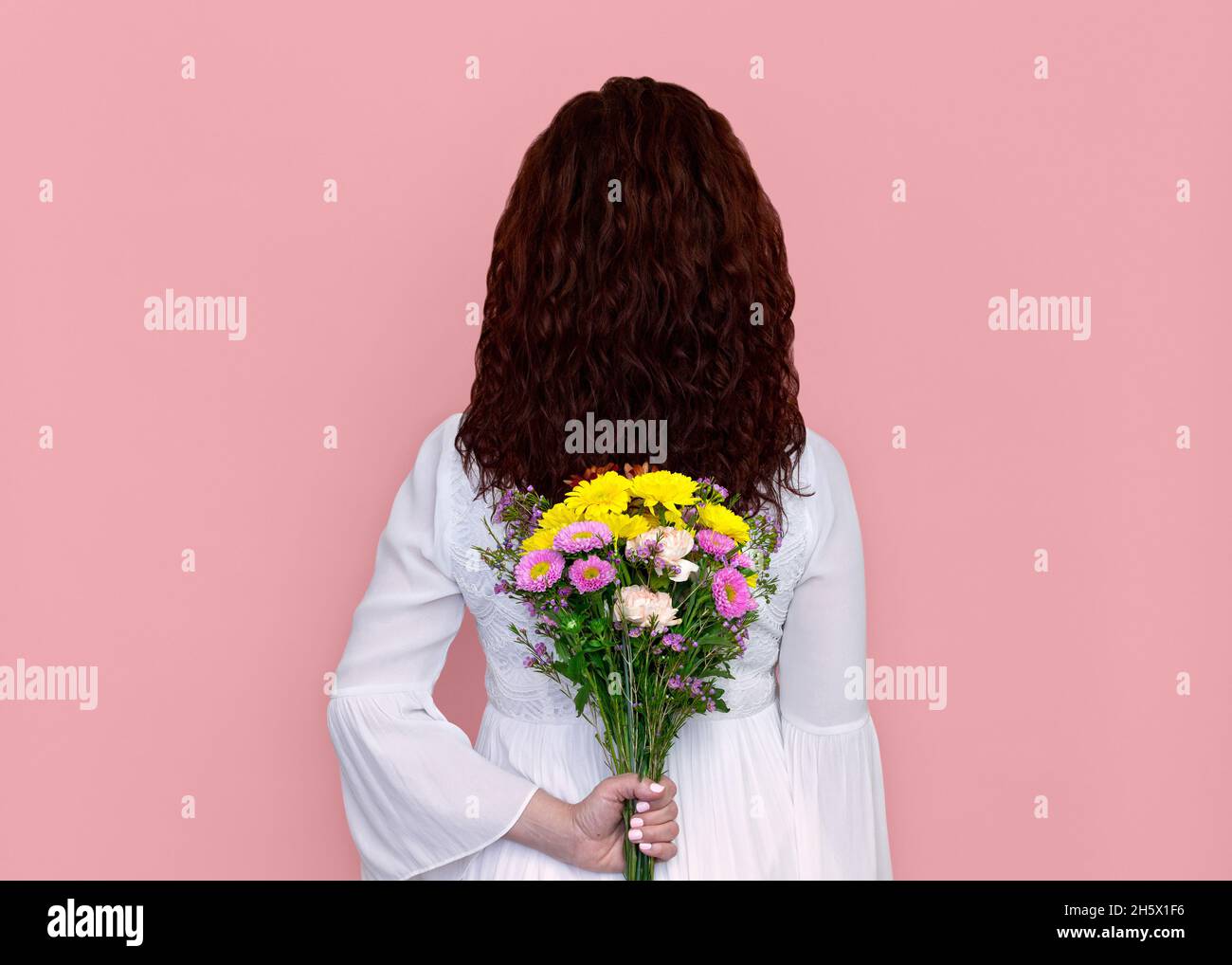 Woman Holding Flowers Behind Her Isolated on Pink Background. Portrait of playful woman in white dress holding Gerbera daisies, carnations, and mums. Stock Photo