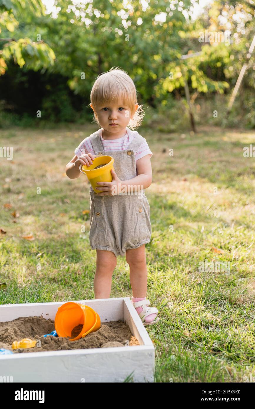 A little girl in a gray jumpsuit with shorts with a yellow toy bucket with sand in her hands is playing in the garden and looking at the camera. There Stock Photo