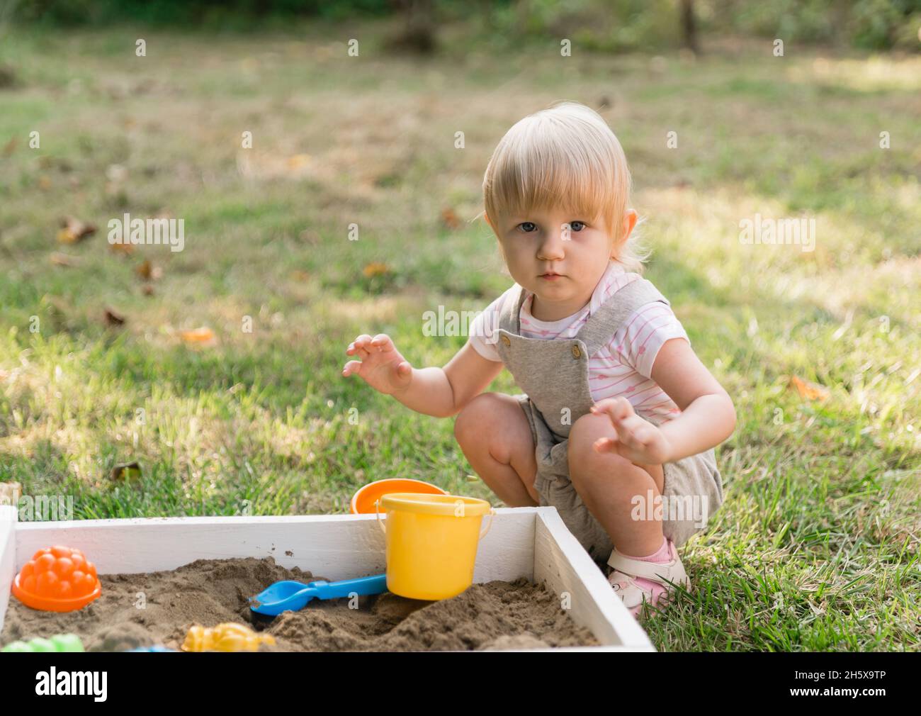 A little beautiful serious girl is sitting in a gray jumpsuit with shorts and playing with sand in the garden. There are toys in the sandbox Stock Photo