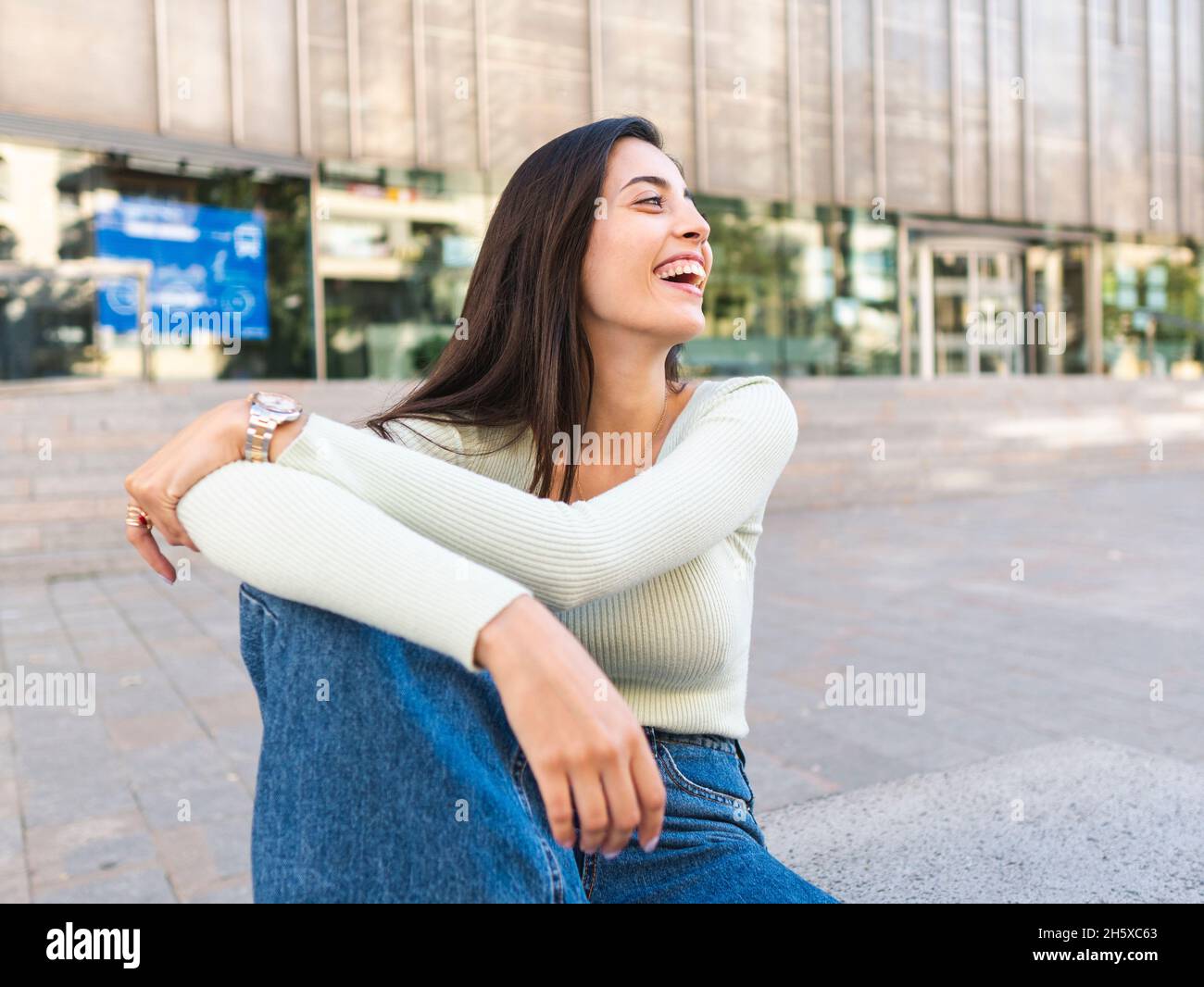 Happy Woman with brown hair in casual clothes sitting on concrete border and embracing knee on city street in daylight Stock Photo