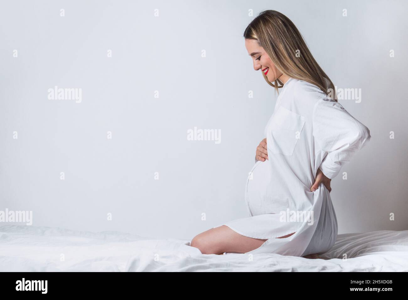 Side view of young gentle female touching tummy while sitting on bed and smiling happily Stock Photo