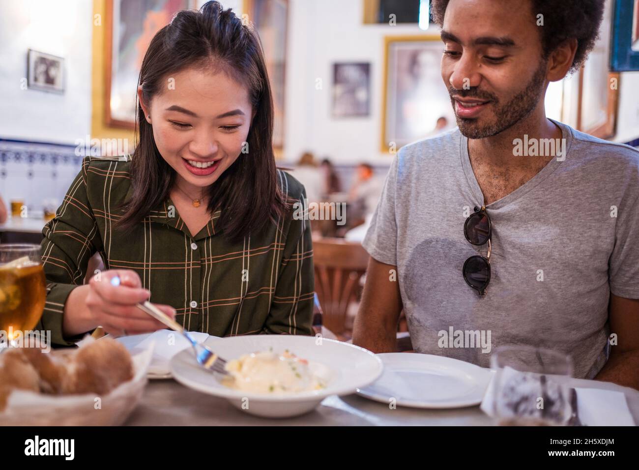 Happy young Asian lady with dark hair in casual clothes smiling while eating delicious salad during lunch with ethnic boyfriend in restaurant Stock Photo