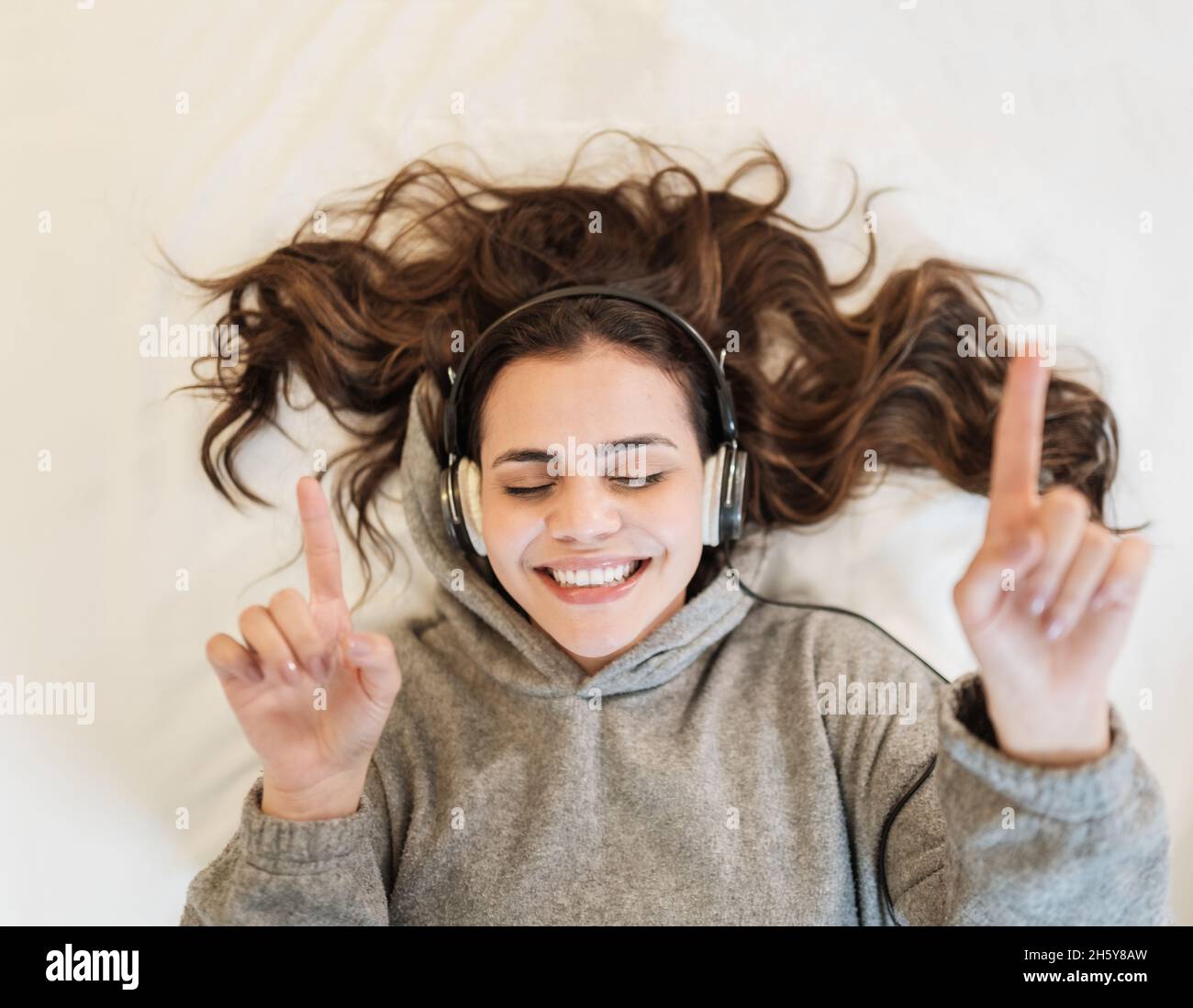 Happy Latin woman wear headphones lying in bed listening music singing