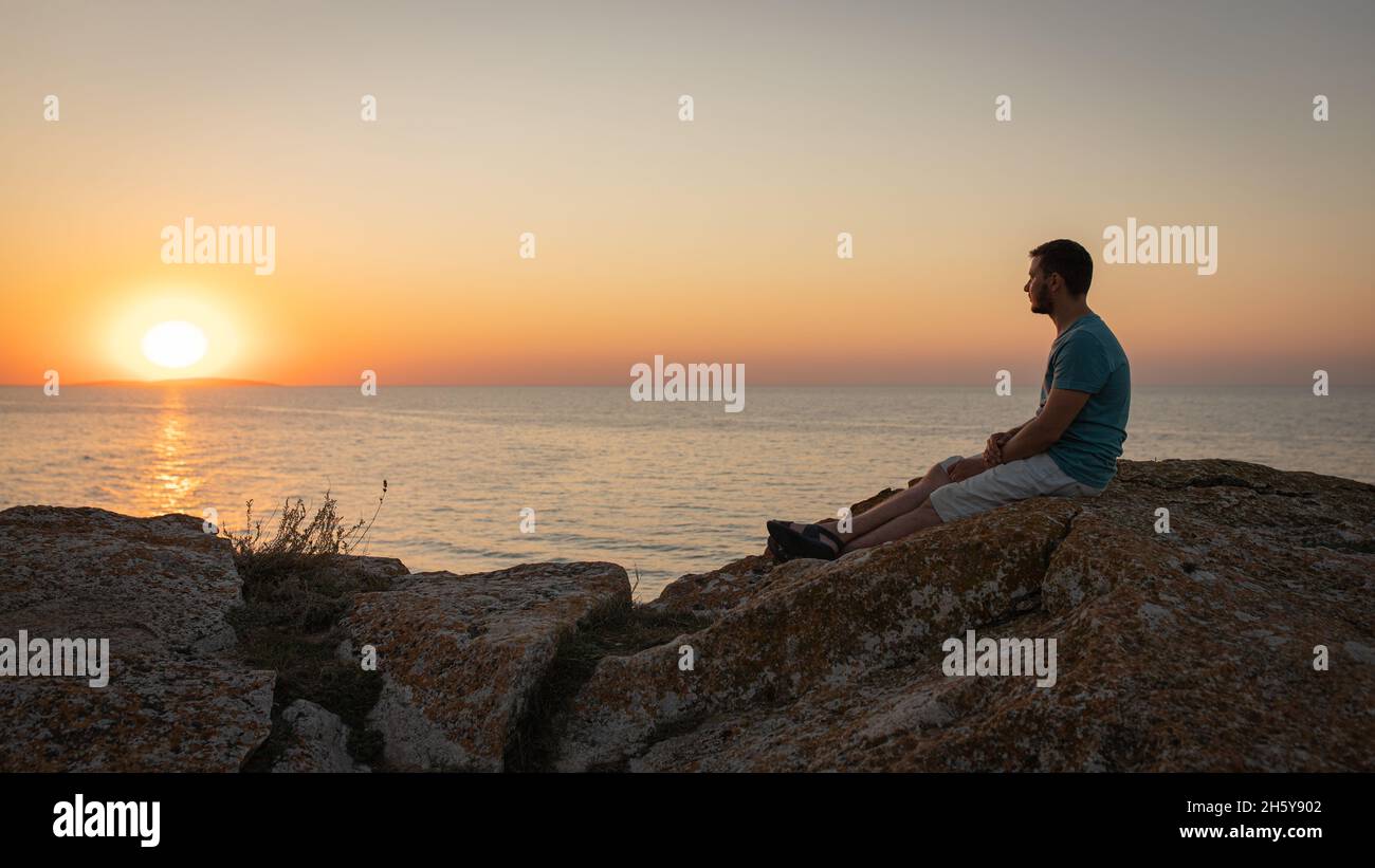 A man enjoys the view of the sunset on the sea, sitting on a rock. Side view, silhouette. Stock Photo
